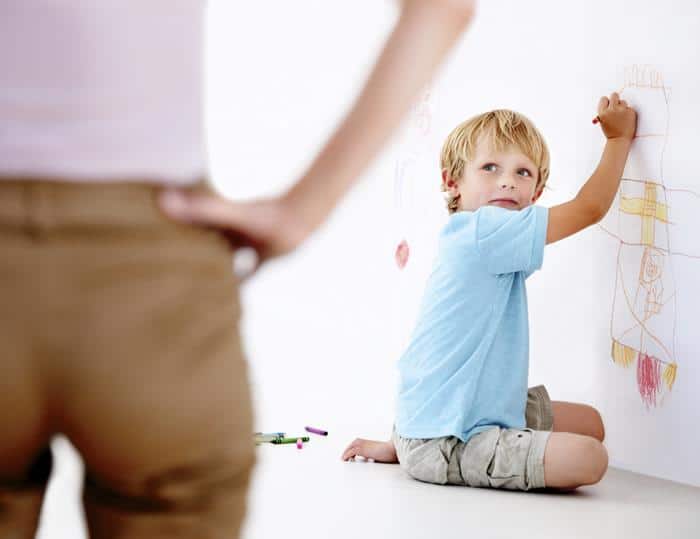 The image shows a boy sitting on the floor and colouring the wall and turned back to see his father watching him with his hand in hip.